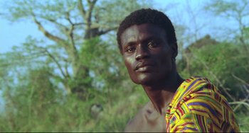 Movie still from “Cobra Verde” (1987), directed by Werner Herzog – A man in a yellow and red striped shirt is standing in front of a tree; Close Up shot, Low angle