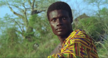 Movie still from “Cobra Verde” (1987), directed by Werner Herzog – A man in a colorful shirt standing in front of a tree; Close Up shot, Low angle