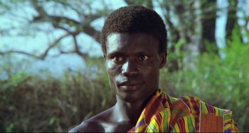 Movie still from “Cobra Verde” (1987), directed by Werner Herzog – A man in a colorful shirt is standing in front of a tree; Close Up shot, Low angle