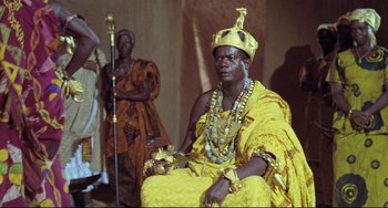 Movie still from “Cobra Verde” (1987), directed by Werner Herzog – A man sitting on top of a chair wearing a crown; Medium shot, Low angle