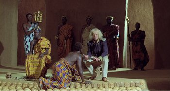 Movie still from “Cobra Verde” (1987), directed by Werner Herzog – A group of men sitting on the ground in front of a building; Wide shot, High angle