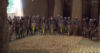 Movie still from “Cobra Verde” (1987), directed by Werner Herzog – A group of men standing next to each other in a room; Wide shot, High angle