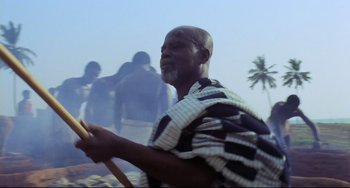 Movie still from “Cobra Verde” (1987), directed by Werner Herzog – An african man holding a wooden stick in his hand; Medium shot, Low angle