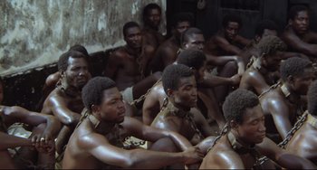Movie still from “Cobra Verde” (1987), directed by Werner Herzog – A group of men chained to each other sitting in a room; Wide shot, High angle