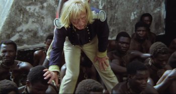 Movie still from “Cobra Verde” (1987), directed by Werner Herzog – An older woman is standing in front of a group of people; Medium shot, High angle