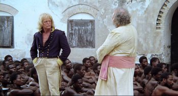 Movie still from “Cobra Verde” (1987), directed by Werner Herzog – Two men are standing next to each other in front of a group of people; Medium shot, Over the shoulder angle