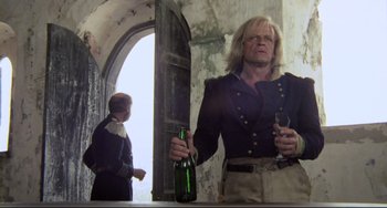 Movie still from “Cobra Verde” (1987), directed by Werner Herzog – A man holding a bottle and a glass in a room; Medium shot, Low angle