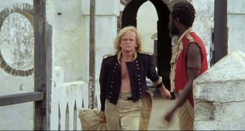 Movie still from “Cobra Verde” (1987), directed by Werner Herzog – A man in a black jacket walking down a street; Medium shot, Low angle
