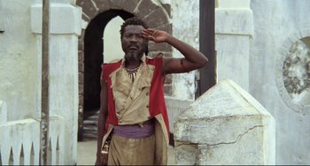Movie still from “Cobra Verde” (1987), directed by Werner Herzog – A man saluting while standing in front of a stone wall; Medium shot, Low angle