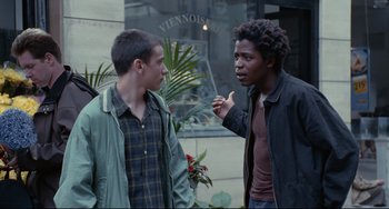 Movie still from “Code Unknown” (2000), directed by Michael Haneke – Two young men standing next to each other on the sidewalk; Medium shot, Over the shoulder angle