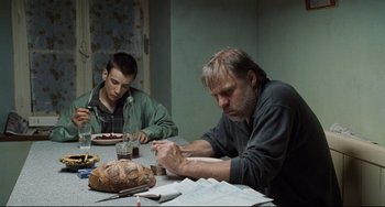 Movie still from “Code Unknown” (2000), directed by Michael Haneke – Two men sitting at a table with a loaf of bread on it; Medium shot, Over the shoulder angle
