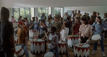 Movie still from “Code Unknown” (2000), directed by Michael Haneke – A group of people standing in a room playing drums; Wide shot, High angle