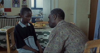 Movie still from “Code Unknown” (2000), directed by Michael Haneke – A man and a boy are sitting at a kitchen table; Medium shot, Over the shoulder angle