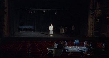 Movie still from “Code Unknown” (2000), directed by Michael Haneke – A woman standing on a stage in front of an audience; Extreme Wide shot, High angle