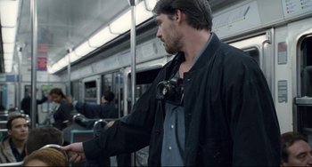 Movie still from “Code Unknown” (2000), directed by Michael Haneke – A man with a camera strapped to his neck is standing on a subway train; Medium shot, Over the shoulder angle