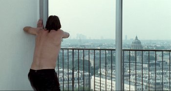 Movie still from “Code Unknown” (2000), directed by Michael Haneke – A man standing in front of a window looking out at a city; Medium shot, Over the shoulder angle