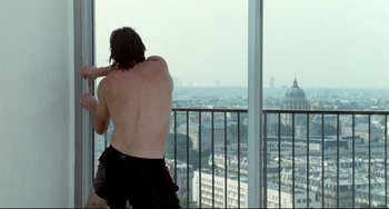 Movie still from “Code Unknown” (2000), directed by Michael Haneke – A man standing in front of a large window with a view of a city; Medium shot, Over the shoulder angle
