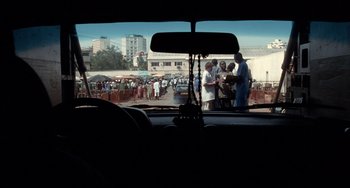 Movie still from “Code Unknown” (2000), directed by Michael Haneke – A group of people standing in a parking lot next to a street; Wide shot, Over the shoulder angle