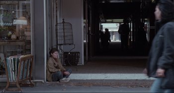 Movie still from “Code Unknown” (2000), directed by Michael Haneke – A person sitting on the ground in front of a building; Wide shot, Over the shoulder angle