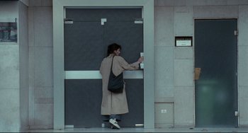 Movie still from “Code Unknown” (2000), directed by Michael Haneke – A woman in a trench coat is standing in front of a door; Wide shot, High angle