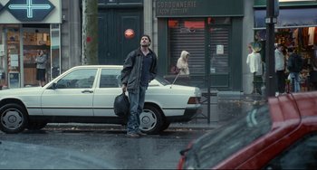 Movie still from “Code Unknown” (2000), directed by Michael Haneke – A man standing on the side of the street holding a bag; Wide shot, Over the shoulder angle