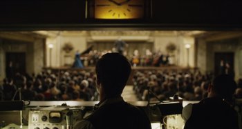 Movie still from “The Death of Stalin” (2017), directed by Armando Iannucci – A man standing in front of an audience in front of a clock; Wide shot, Over the shoulder angle