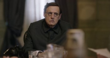 Movie still from “The Death of Stalin” (2017), directed by Armando Iannucci – An older man sitting at a table in front of glasses; Close Up shot, Over the shoulder angle