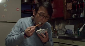 Movie still from “Cold Fish” (2010), directed by Sion Sono – A man in a blue sweater is eating food from a bowl; Close Up shot, Overhead angle