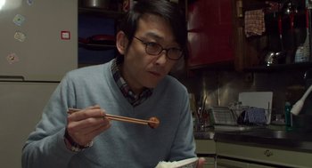 Movie still from “Cold Fish” (2010), directed by Sion Sono – A man in glasses eating food with chopsticks in a kitchen; Close Up shot, High angle
