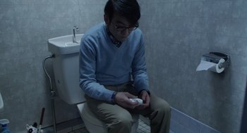 Movie still from “Cold Fish” (2010), directed by Sion Sono – A man sitting on a toilet in a bathroom holding a nintendo wii game controller; Medium shot, Low angle