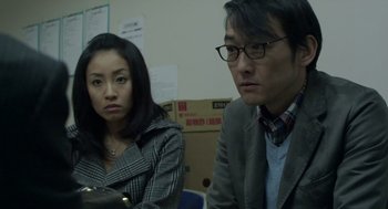 Movie still from “Cold Fish” (2010), directed by Sion Sono – A man and a woman sitting next to each other in front of boxes; Close Up shot, High angle