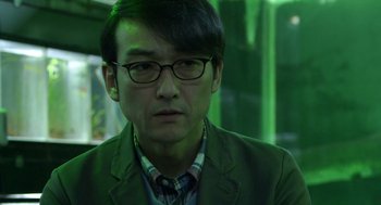 Movie still from “Cold Fish” (2010), directed by Sion Sono – A man in a green jacket wearing glasses; Close Up shot, Low angle