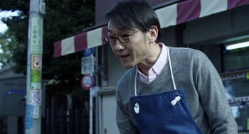 Movie still from “Cold Fish” (2010), directed by Sion Sono – A man wearing an apron and glasses is standing in front of a building; Medium shot, Low angle