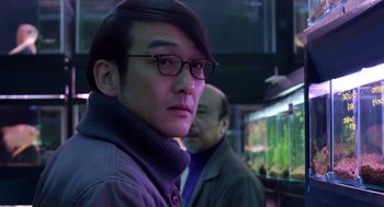 Movie still from “Cold Fish” (2010), directed by Sion Sono – A man wearing glasses and a jacket is looking at the camera; Close Up shot, Over the shoulder angle