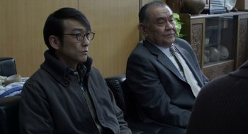 Movie still from “Cold Fish” (2010), directed by Sion Sono – Two men in suits sitting in front of each other; Medium shot, Over the shoulder angle