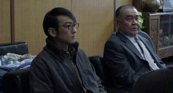 Movie still from “Cold Fish” (2010), directed by Sion Sono – Two men sitting next to each other in front of a wooden wall; Medium shot, Low angle