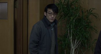 Movie still from “Cold Fish” (2010), directed by Sion Sono – A man in a jacket standing in front of a plant; Medium shot, High angle