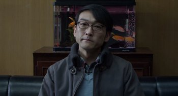 Movie still from “Cold Fish” (2010), directed by Sion Sono – A man with glasses is in front of a fish tank; Close Up shot, Over the shoulder angle