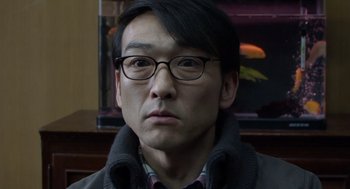 Movie still from “Cold Fish” (2010), directed by Sion Sono – A person wearing glasses and a jacket; Close Up shot, Over the shoulder angle