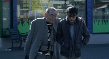 Movie still from “Cold Fish” (2010), directed by Sion Sono – Two men walking down a street with their arms around one another; Medium shot, Low angle