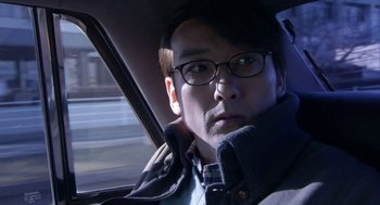 Movie still from “Cold Fish” (2010), directed by Sion Sono – A man with glasses sitting in the back seat of a car; Close Up shot, Over the shoulder angle