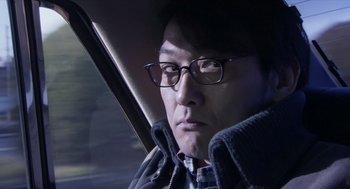 Movie still from “Cold Fish” (2010), directed by Sion Sono – A man wearing glasses sitting in the back of a car; Close Up shot, Low angle