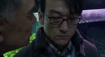 Movie still from “Cold Fish” (2010), directed by Sion Sono – A person wearing glasses and a jacket; Close Up shot, Low angle