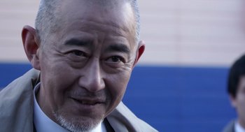Movie still from “Cold Fish” (2010), directed by Sion Sono – An older man with a goatee is smiling for the camera; Close Up shot, Low angle