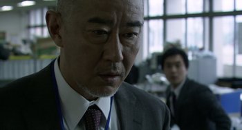 Movie still from “Cold Fish” (2010), directed by Sion Sono – An older man in a suit and tie looking down; Close Up shot, Over the shoulder angle