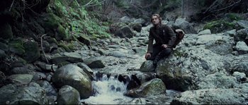 Movie still from “Cold Mountain” (2003), directed by Anthony Minghella – A man sitting on top of a rock near a stream; Wide shot, Low angle