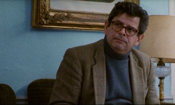 Movie still from “Cold Water” (1994), directed by Olivier Assayas – A man wearing glasses and a jacket sitting in front of a painting; Close Up shot, Over the shoulder angle