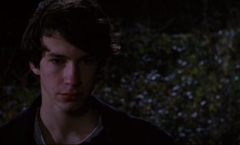 Movie still from “Cold Water” (1994), directed by Olivier Assayas – A young man is looking at the camera while standing in front of a bush; Close Up shot, Over the shoulder angle