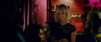 Movie still from “Collide” (2016), directed by Eran Creevy – A woman smiles at the camera while two other women sit at a table; Close Up shot, Over the shoulder angle