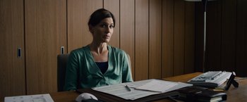Movie still from “Collide” (2016), directed by Eran Creevy – A woman sitting in front of papers on top of a table; Medium shot, Over the shoulder angle